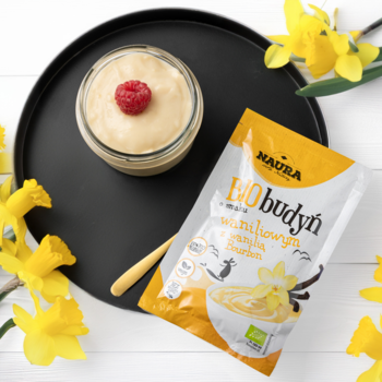 Widok z góry: Hauba BIO budyń waniliowy bez cukru w żółtej torebce obok gotowego deseru na czarnej tacy.