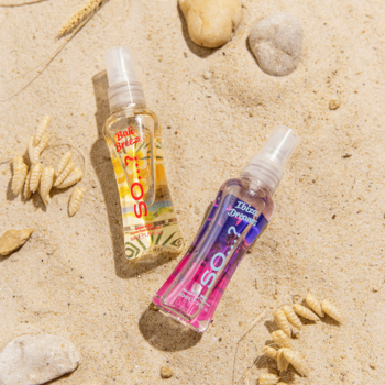 Dwie mgiełki do ciała So...? (Bali Breeze różowa, Pink Gold bursztynowa) na piaszczystej plaży z kamykami. Widok z góry.