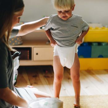 Chłopiec w białych pieluchomajtkach Bambiboo z bambusowego włókna, ubiera się z pomocą mamy w pokoju.