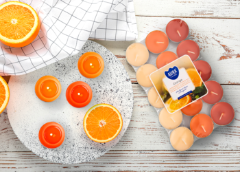 Pomarańczowe świece zapachowe typu tealight, ułożone z plastrami pomarańczy i opakowaniem na białym, drewnianym stole.