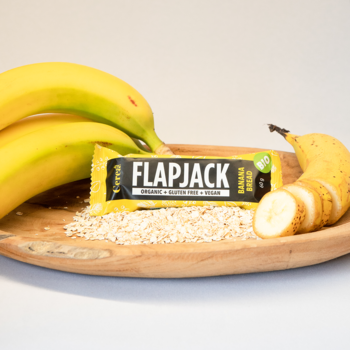 Ekologiczny baton owsiany Flapjack Bananowy Chlebek w żółtej folii, z bananami i owsem na drewnianej tacy.