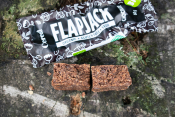 Flapjack baton owsiany wegański z orzechami nerkowca i czekoladą, przełamany na pół, obok opakowania na drewnie.