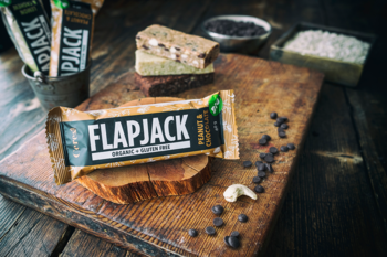 Pieczony baton owsiany Flapjack z orzeszkami ziemnymi i czekoladą na drewnianej desce, obok rozsypane składniki.