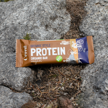 Ekologiczny baton proteinowy Cerea Brownie Style BIO, brązowo-pomarańczowe opakowanie, leży na szarych skałach.