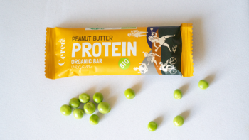 Żółty, organiczny baton proteinowy 'Peanut Butter Protein Bar' na białym tle, obok zielone groszki.