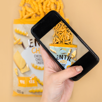 Ręka robiąca smartfonem zdjęcie otwartej torebki Lentil Chips o smaku sera cheddar.
