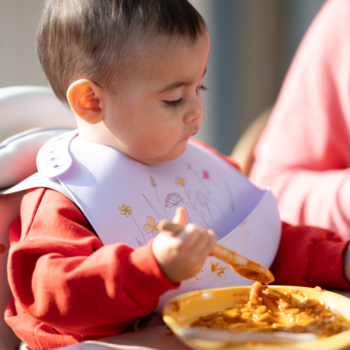 Dziecko w fioletowym śliniaku je gulasz Bobovita z żółtej miseczki łyżeczką, widok z boku, obiad.