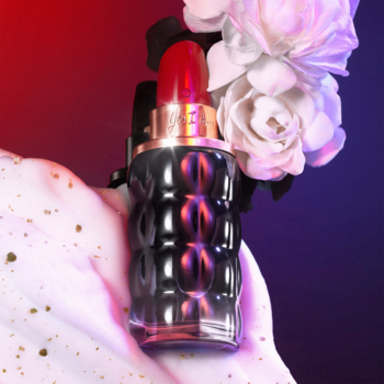 Cacharel Yes I Am, butelka perfum o ciemnej, prążkowanej teksturze, z czerwoną "pomadką", trzymana wśród kwiatów.