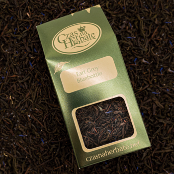 Herbata Earl Grey Bluebottle w zielonej paczce na rozsypanych czarnych liściach z niebieskimi płatkami, widok z góry.