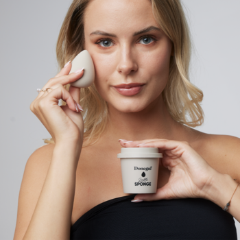 Kobieta trzyma beżową gąbkę Blending Sponge do podkładu przy twarzy i pasujące etui w kształcie kubka.