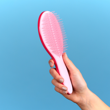 Różowa szczotka Tangle Teezer z rączką, uniesiona w dłoni, z widocznymi elastycznymi ząbkami na niebieskim tle.