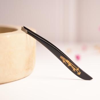 Czarna skośna pęseta do brwi ze stali nierdzewnej, ze złotym wzorem, leży pod kątem na jasnym tle.