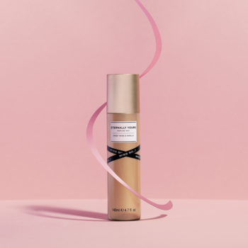 Mgiełka perfumowana Eternally Yours w złotej butelce z czarną kokardą, stojąca na różowym tle z różową wstążką.