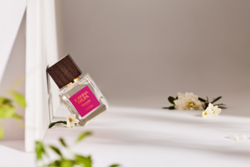 Perfumy Secret Sense w przezroczystej butelce z różową etykietą, skośnie ustawione na białym tle z kwiatami.