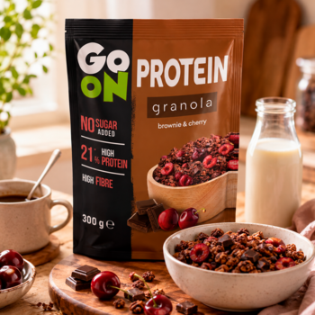 Brązowo-zielona torebka Go On Protein Granola Brownie Wiśnia 300g, bez cukru, na stole obok miski z granolą.