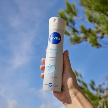 Biały dezodorant NIVEA Fresh Natural spray trzymany w dłoni na tle błękitnego nieba z zielonymi liśćmi drzew.