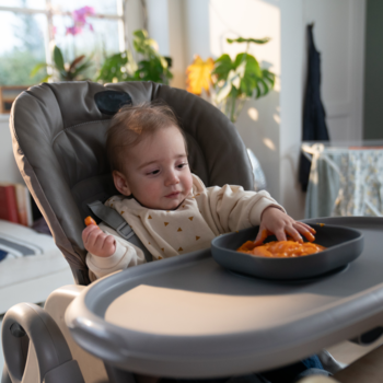 Dziecko w krzesełku do karmienia je zupę jarzynową Bobovita, brudząc się pomarańczowym jedzeniem w miseczce.