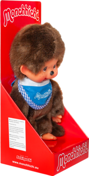Brązowa pluszowa lalka Monchhichi w niebieskim śliniaku w kratkę, siedzi w czerwonym opakowaniu prezentacyjnym.