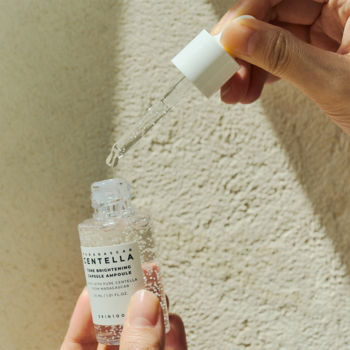 SKIN1004 serum rozjaśniające z wąkrotą azjatycką, dłoń trzyma butelkę i zakraplacz z kroplą, na jasnym tle.