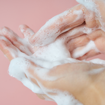 Dłonie pokryte gęstą pianką do mycia twarzy z wąkrotą azjatycką, na jasnoróżowym tle.