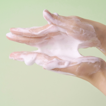 Zbliżenie na dłonie, gęsta pianka do mycia twarzy SKIN1004 Madagascar Centella Tea-Trica na jasnym zielonym tle.