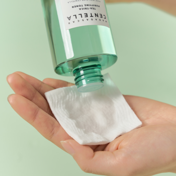 Tonik Centella Tea Tree w zielonej butelce nalewany na biały płatek kosmetyczny w dłoni, zbliżenie.