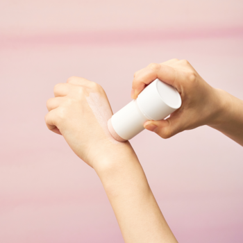 Maska z glinką w białym sztyfcie, aplikowana na dłoń z widocznym śladem produktu, na różowym tle.