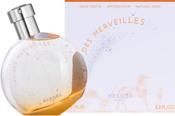 Hermes Eau des Merveilles, okrągła butelka perfum z pomarańczowym płynem i brokatem, przed białym pudełkiem.