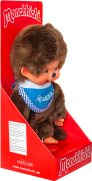 Brązowa pluszowa lalka Monchhichi w niebieskim śliniaku w kratkę, siedzi w czerwonym opakowaniu prezentacyjnym.