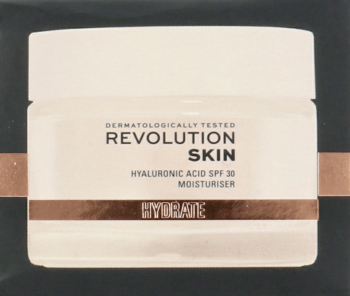 Biały słoiczek kremu Revolution Skin Hyaluronic Acid SPF 30 z miedzianą opaską, widok z przodu przez ciemnoszare pudełko.